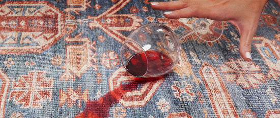 Hand with white painted nails reaching down to a spilled wine glass with red wine soaking into a machine washable rug.
